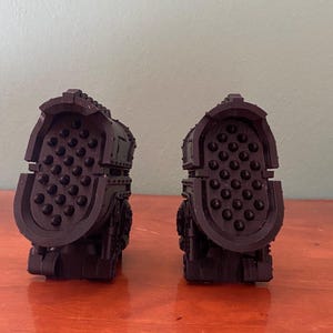 Op de afbeelding: Twee donkerpaarse, gegoten kunststof onderdelen met een opvallende ovale voorzijde. Elke voorzijde is bedekt met een raster van kleine, verhoogde ronde noppen. De structuren hebben een mechanisch en industrieel uiterlijk met panelen.