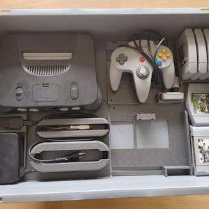 Könnte beinhalten: Eine graue Nintendo 64 Konsole mit passendem Controller und mehreren Spielkassetten, ordentlich in einem grauen Aufbewahrungsbehälter verstaut. Konsole und Zubehör sind in gutem Zustand, was auf sorgfältige Nutzung und Lagerung hindeutet.