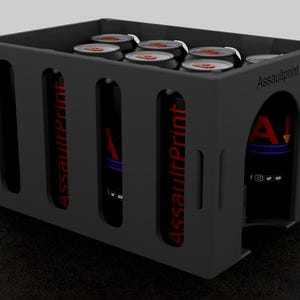 Könnte beinhalten: Eine schwarze, rechteckige Transportkiste mit ausgeschnittenen Griffen und dem roten Text "AssaultPrint". Die Kiste enthält mehrere Dosen mit roten und weißen Deckeln. Das Logo "A!" ist auf der Seite sichtbar.
