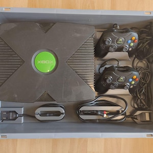 Könnte beinhalten: Eine schwarze Xbox-Konsole mit grünem Power-Knopf und zwei schwarzen Controllern befinden sich in einer grauen Aufbewahrungsbox. Kabel und Anschlüsse sind ebenfalls sichtbar. Die Xbox ist eine klassische Spielekonsole.