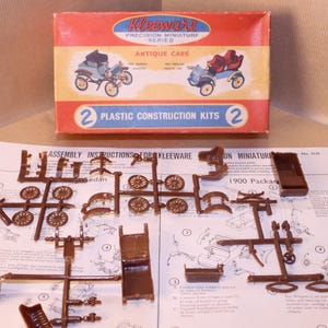 May include: A vintage model car kit with two plastic construction kits. The box features a red, white, and blue colour scheme with the text "Kleeware Precision Miniature Series Antique Cars". The instructions include a diagram of the car parts and the text "Assembly Instructions for Kleeware On Miniature 1900 Packag".