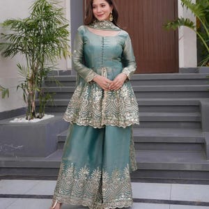 May include: A teal-colored, long-sleeved outfit with intricate gold embroidery. The ensemble includes a tunic-style top with a peplum and wide-leg pants. The outfit is accessorized with matching jewelry.
