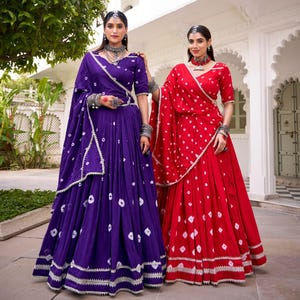 May include: Two women in traditional Indian outfits. One wears a deep purple outfit with white dot patterns, a matching shawl, and silver jewellery. The other wears a red outfit with white dot patterns, a matching shawl, and silver jewellery. Both outfits have intricate silver trim.