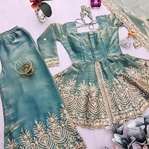 May include: A teal-colored, embroidered formal outfit, including a top, pants, and a scarf. The top features a square neckline and long sleeves. The outfit is embellished with gold embroidery and sequins. A small toy car and decorative flowers are also present.