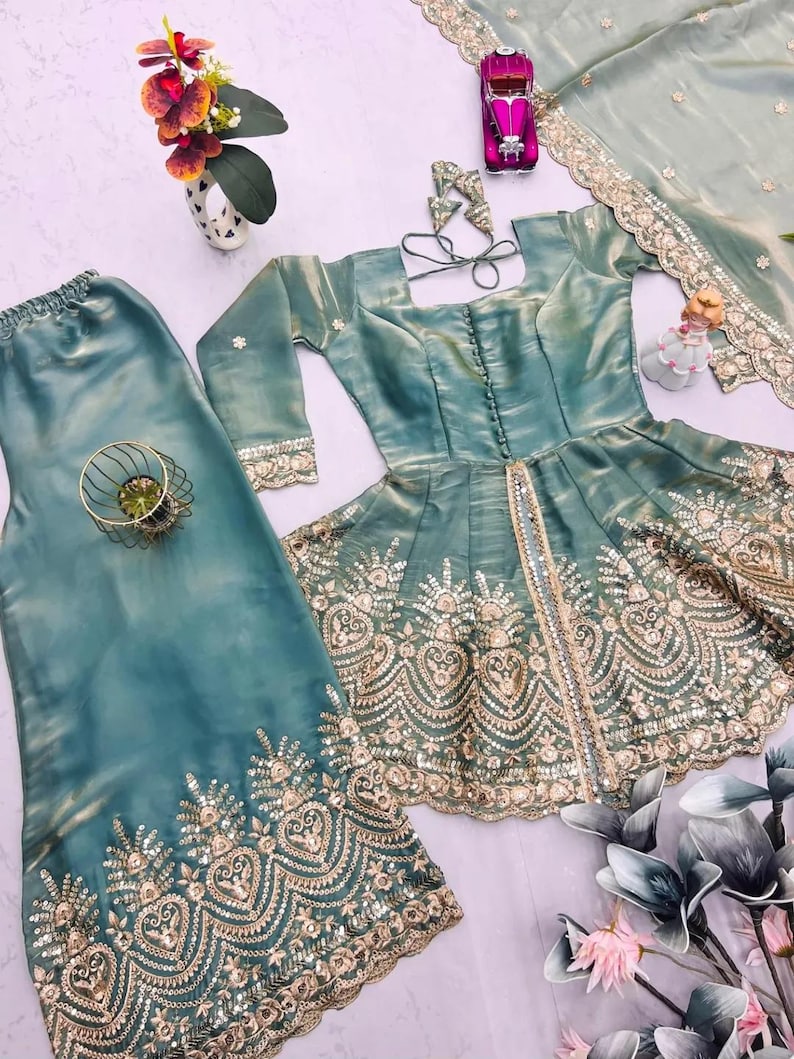 May include: A teal-colored, embroidered formal outfit, including a top, pants, and a scarf. The top features a square neckline and long sleeves. The outfit is embellished with gold embroidery and sequins. A small toy car and decorative flowers are also present.