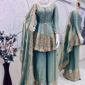 May include: A teal-colored formal outfit displayed on mannequins. The ensemble includes a long-sleeved top with intricate gold embroidery and matching wide-leg pants. A sheer scarf with gold trim drapes over the outfit. A gold necklace is visible.