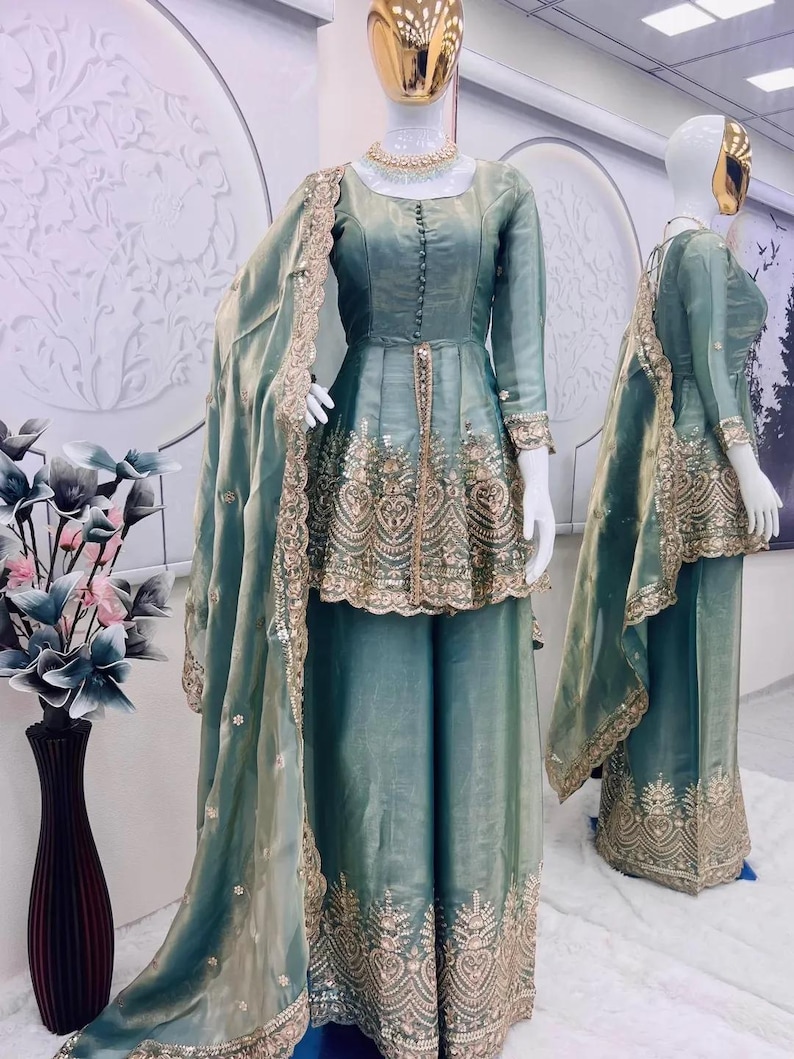 May include: A teal-colored formal outfit displayed on mannequins. The ensemble includes a long-sleeved top with intricate gold embroidery and matching wide-leg pants. A sheer scarf with gold trim drapes over the outfit. A gold necklace is visible.