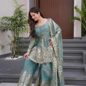 May include: A teal-colored, embroidered formal outfit. The ensemble includes a long-sleeved top with intricate gold embroidery, wide-leg pants, and a matching scarf. The outfit is worn with gold earrings and sandals.