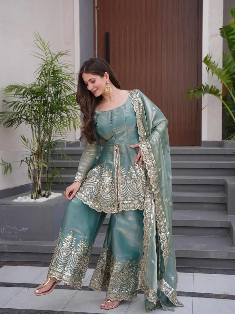 May include: A teal-colored, embroidered formal outfit. The ensemble includes a long-sleeved top with intricate gold embroidery, wide-leg pants, and a matching scarf. The outfit is worn with gold earrings and sandals.