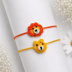 May include: Two handmade crochet bracelets featuring animal motifs. The top bracelet has an orange lion face with a yellow center and white muzzle. The bottom bracelet features a yellow bear face with a white muzzle. Both have thin cord bands.