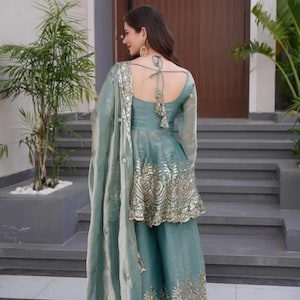May include: A teal-colored, embroidered Indian suit with a matching dupatta. The suit features a peplum top with gold embellishments and wide-leg pants. The dupatta has a delicate, embroidered border. The outfit is worn with gold earrings.