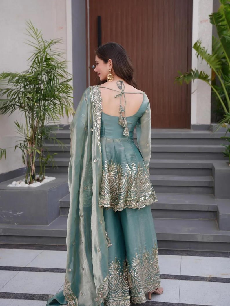 May include: A teal-colored, embroidered Indian suit with a matching dupatta. The suit features a peplum top with gold embellishments and wide-leg pants. The dupatta has a delicate, embroidered border. The outfit is worn with gold earrings.