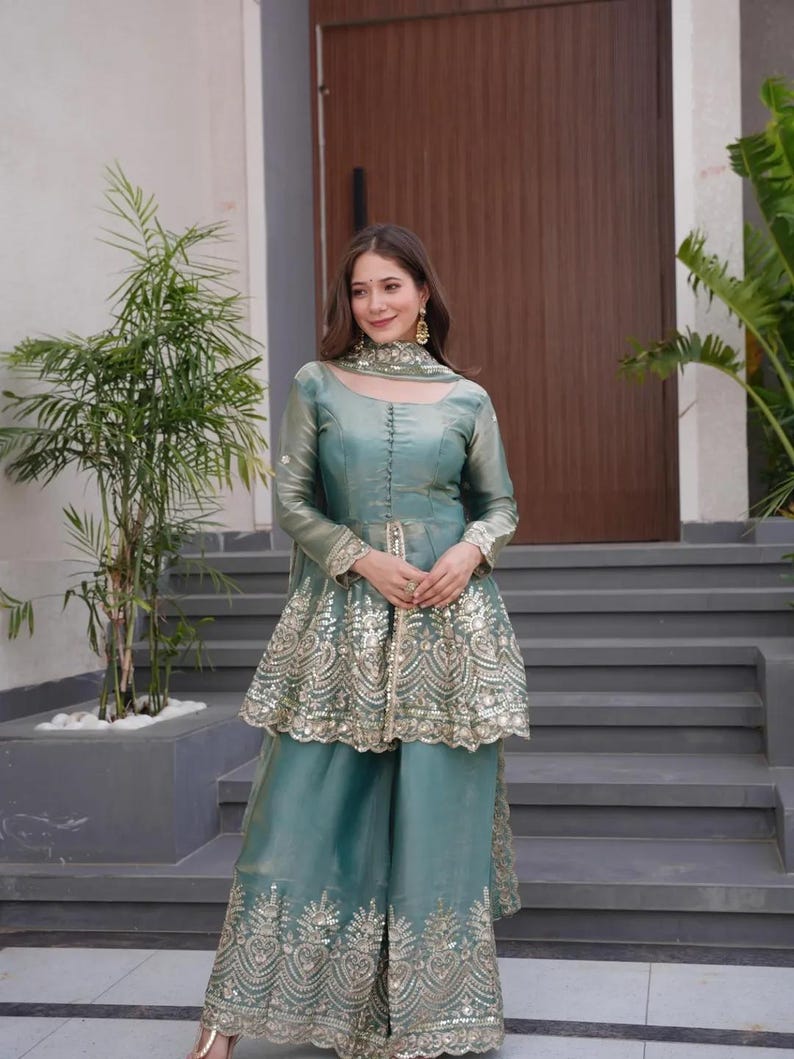 May include: A teal-colored, long-sleeved outfit with intricate gold embroidery. The ensemble includes a tunic-style top with a peplum and wide-leg pants. The outfit is accessorized with matching jewelry.