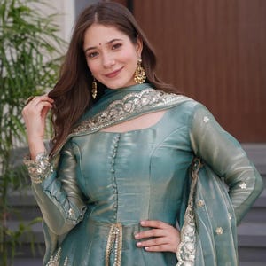 May include: A teal-colored formal dress with long sleeves and gold embellishments. The dress features a button-down bodice and a flowing skirt. The matching shawl is adorned with intricate gold embroidery. The woman is wearing gold earrings.