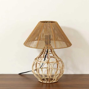 May include: A decorative table lamp with a woven rattan base and a conical shade. The shade is made of tightly wound natural-colored twine. The lamp sits on a dark wooden surface against a white wall.