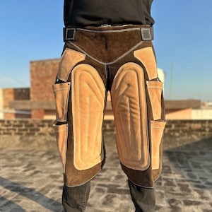 以下が含まれることがあります： 茶色の革製の作業用ズボンで、膝当てと工具ポケットが付いています。ズボンは茶色のスエードのウエストバンドと黒いステッチが付いています。