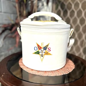 Puede incluir: Una bolsa nevera blanca, cilíndrica, con cierre de cremallera y asa superior. La bolsa presenta un emblema de estrella bordada de colores con las letras O, E y S. Tiene una correa para el hombro y está sobre una alfombrilla decorativa.