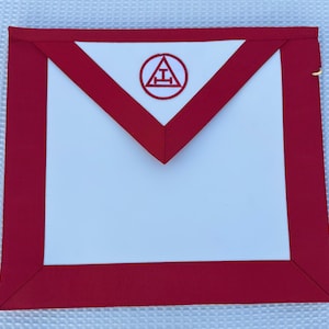 May include: A red and white Masonic apron. The apron has a white center with a red border and a red triangle at the top. A red circle with a symbol is in the center of the triangle. The apron is rectangular in shape.