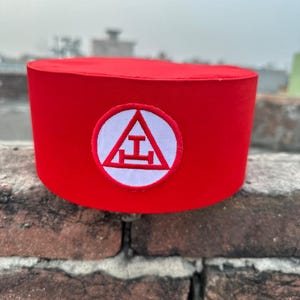 May include: A red felt Masonic hat with a white embroidered emblem featuring a triangle with a letter "T" inside and a square below it.