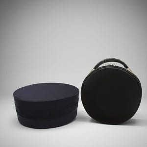 Op de afbeelding: Twee stuks bagage. Een marineblauwe hoedendoos met een zwarte band en een zwarte ronde koffer met een handvat. De hoedendoos is ovaal en de koffer is rond. Beide staan op een witte achtergrond.