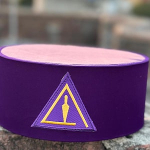 Puede incluir: Un sombrero de terciopelo morado con un triángulo bordado en oro y una figura estilizada en su interior. El triángulo tiene un borde amarillo.