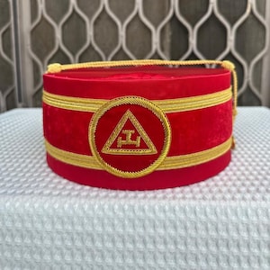 May include: A red velvet hat with gold trim and a gold emblem. The hat is cylindrical with a flat top and bottom. The emblem is a gold triangle with a symbol inside a gold circle.
