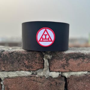 Puede incluir: Collar masónico negro con un emblema bordado en rojo y blanco que representa un triángulo con una letra 'I' y una línea horizontal.