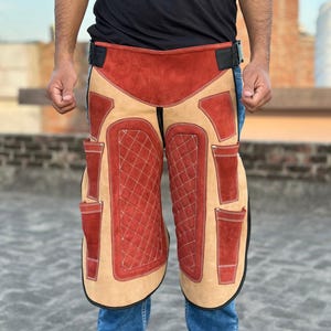 May include: A red and tan leather work apron with multiple pockets and a quilted design. The apron is being worn by a person wearing blue jeans.