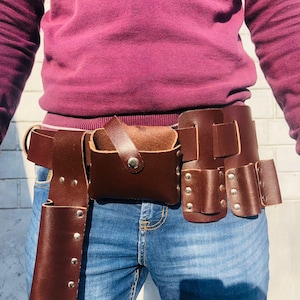 May include: A brown leather tool belt with multiple pockets and loops. The belt is worn over blue jeans.