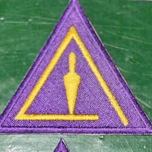 May include: Purple embroidered patch in a triangular shape with a yellow design. The design features a yellow triangle outline and a yellow shape in the center. The patch is on a green surface.