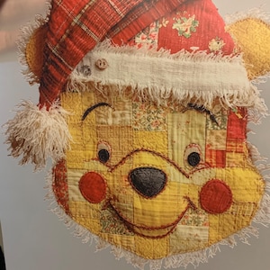 May include: A handcrafted, quilted Winnie the Pooh face wearing a Santa hat. The hat is red plaid and floral fabric with a white trim. The bear's face is yellow patchwork with red cheeks and a black nose. The edges are fringed.