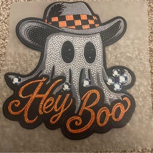 Può includere: Una toppa da stirare nera e arancione con un fantasma che indossa un cappello a scacchi e il testo "Hey Boo".