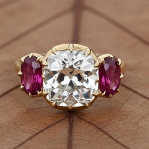 Può includere: Un anello d'oro con una grande pietra preziosa trasparente a taglio quadrato affiancata da due pietre preziose rosse scure a taglio ovale. L'anello è su uno sfondo marrone.