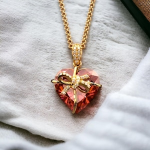 May include: A gold chain necklace with a pink heart-shaped pendant. The pendant is decorated with a bow and small clear stones.