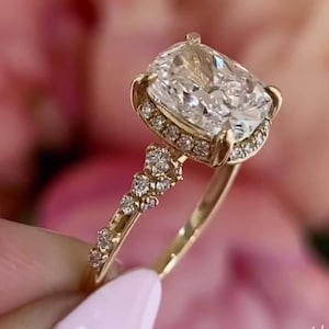 May include: A gold ring featuring a large, square-cut diamond center stone. The ring has a halo of smaller diamonds around the center stone and along the band. The ring is held up against a soft pink background.