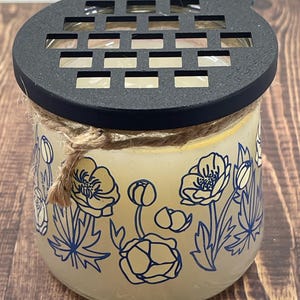 May include: A clear glass jar candle with a black metal lid and a floral design. The jar has a white background with blue floral patterns and a yellow wax inside.