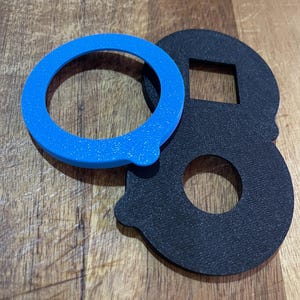 Op de afbeelding: Een blauw, cirkelvormig object met een klein lipje ligt naast een zwart, schedelvormig item met een vierkante uitsparing. De items liggen op een houten oppervlak. Het blauwe object is een ring en het zwarte item heeft een cirkelvormige opening.