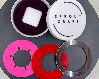 Könnte beinhalten: Ein weißer Behälter mit einem schwarzen Deckel, auf dem "SPROUT CRAFT" steht, und einer violetten Flüssigkeit im Inneren. Es gibt drei runde Scheiben, eine rosa mit einem Sonnenmotiv, eine rote und eine silberne.