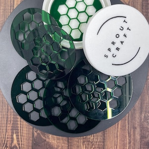 May include: A collection of green, honeycomb-patterned coasters and a white circular item with the text "SPROUT CRAFT" in black. The coasters are arranged on a dark gray surface, with a wooden background.