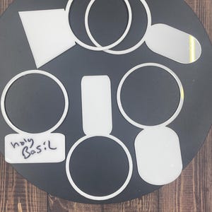 Può includere: Una collezione di segnaposto per piante in acrilico bianco di varie forme, tra cui cerchi, rettangoli e un trapezio. Un segnaposto presenta il testo scritto a mano "holy Basil" in inchiostro nero. Questi oggetti piatti e lisci sono disposti in cerchio.