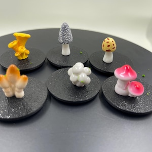 Puede incluir: Siete figuritas de setas en miniatura sobre bases circulares negras. Las setas son de varios colores y formas, incluyendo amarillo, blanco, marrón y rosa.