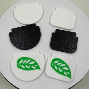 May include: Four different designs of white and black plastic pot covers. Two covers are plain white, two are plain black, and two have a green leaf design.