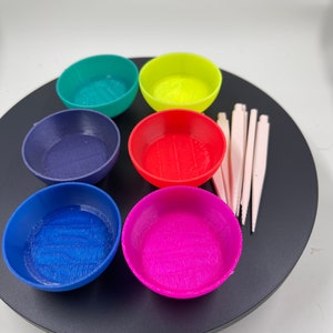 May include: Six small, colorful bowls arranged in a circle on a black surface. The bowls are green, yellow, blue, red, pink, and purple. There are also several wooden sticks in the image.