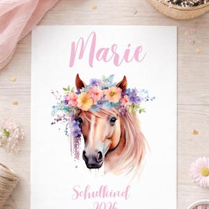 Op de afbeelding: Een aquarelprint met een paardenhoofd versierd met een bloemenkrans. De naam "Marie" staat in roze schrift boven het paard, met "Schulkind 2026" eronder. Het paard heeft een lichtbruine vacht en een kleurrijke bloemenkrans.
