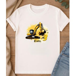 Puede incluir: Camiseta blanca con un diseño de excavadora amarilla y negra, el número 3 y el nombre "EMIL". El gráfico está sobre un fondo amarillo. La camiseta está hecha de un material suave.