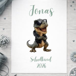 May include: A white rectangular print featuring a green-toned illustration of a Tyrannosaurus Rex wearing black sunglasses and a black hoodie with arms crossed. The text Jonas appears above the dinosaur, and Schulkind 2026 is printed below.