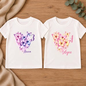 May include: Two white short-sleeved t-shirts featuring butterfly graphics arranged in heart shapes. The left shirt displays purple butterflies, the number 3, and the name Anna. The right shirt displays yellow and pink butterflies, the number 4, and the name Selyne.