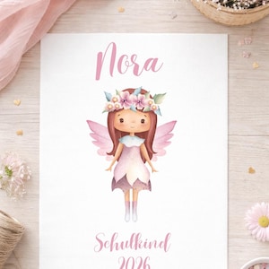 Peut inclure: Une impression rectangulaire blanche présentant une illustration à l'aquarelle d'une fée aux cheveux bruns, avec une couronne de fleurs et des ailes roses. Le texte Nora est imprimé au-dessus et Schulkind 2026 est imprimé en rose en dessous.