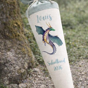 May include: A conical fabric gift cone featuring a light beige body with a sage green fabric top tied with a cord. The cone displays a colorful illustration of a dragon, the name Louis, and the text Schulkind 2026 in a blue script font.