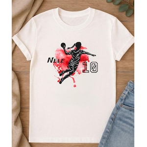 May include: White t-shirt featuring a black silhouette of a person playing handball, with the name "Nele" and the number "10" in black. The design includes a red watercolor splash.
